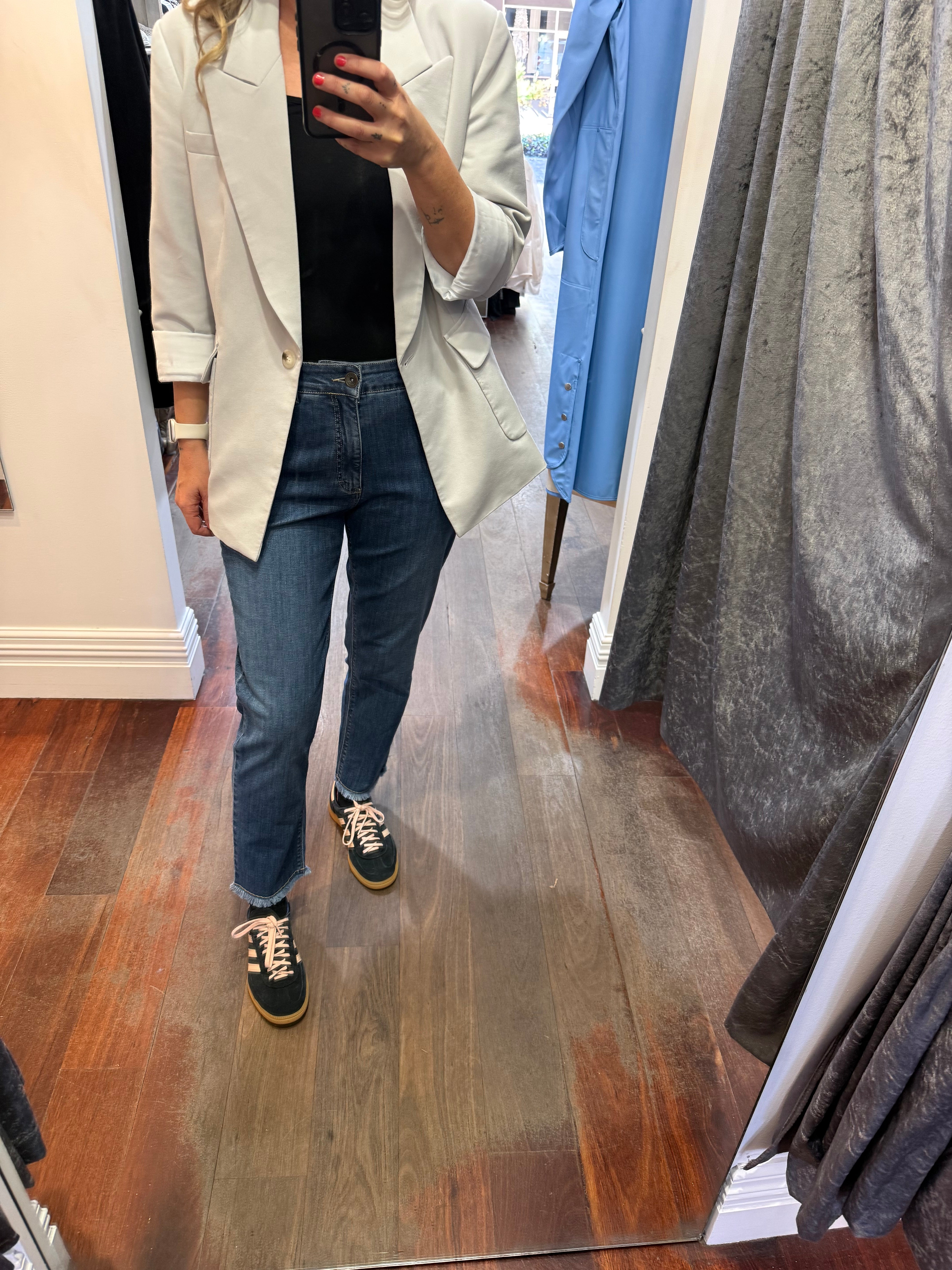 Person wearing a light gray blazer, black top, blue jeans, and black sneakers in a fitting room.